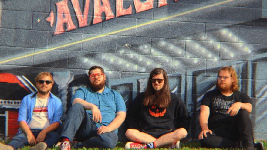  Four men sit on the grass with their backs against a cinder-block wall decorated with street art.