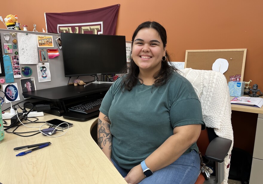 A young woman in a green shirt and jeans sits at a desk.