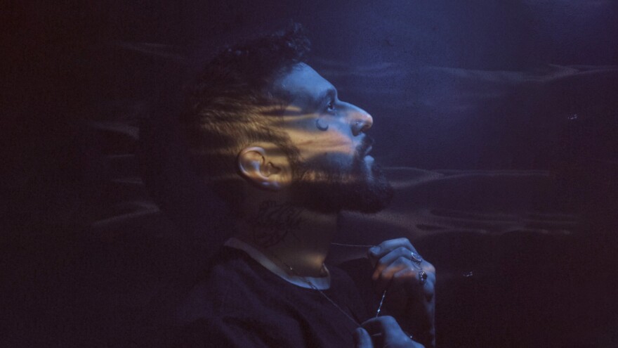 A side profile of a man with a beard and a crescent-moon tattoo near his eye set against a dark, moody background.