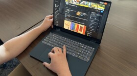 A black laptop sits on a dark brown, woodgrain table. On its screen is the homepage of r/columbiamo, Columbia's Reddit page. Someone largely offscreen is using the laptop, with their left hand resting next to the left side of the screen and their right hand using the trackpad. 