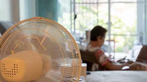 Close-up of a white electric fan in a cozy living room, cooling the space while an elderly woman relaxes in the background. Concept of summer heat, home comfort, and elderly care indoors.