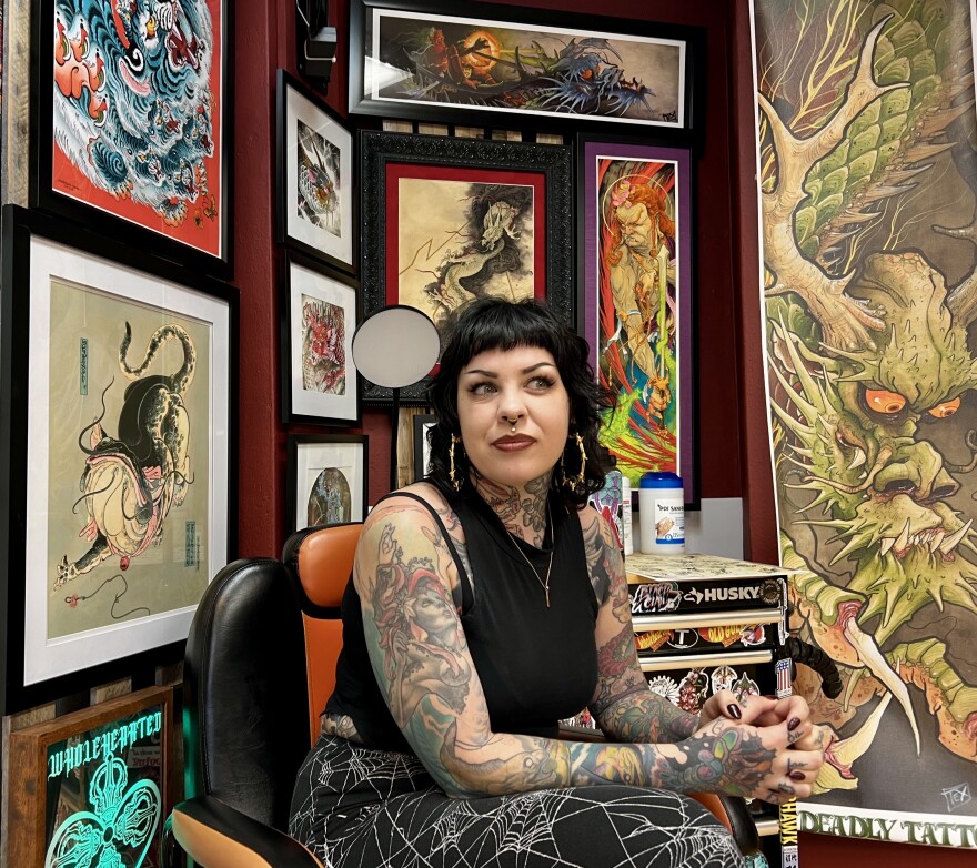 A woman with tattoo sleeves on both arms sits in a chair in front of a wall full of framed art. 
