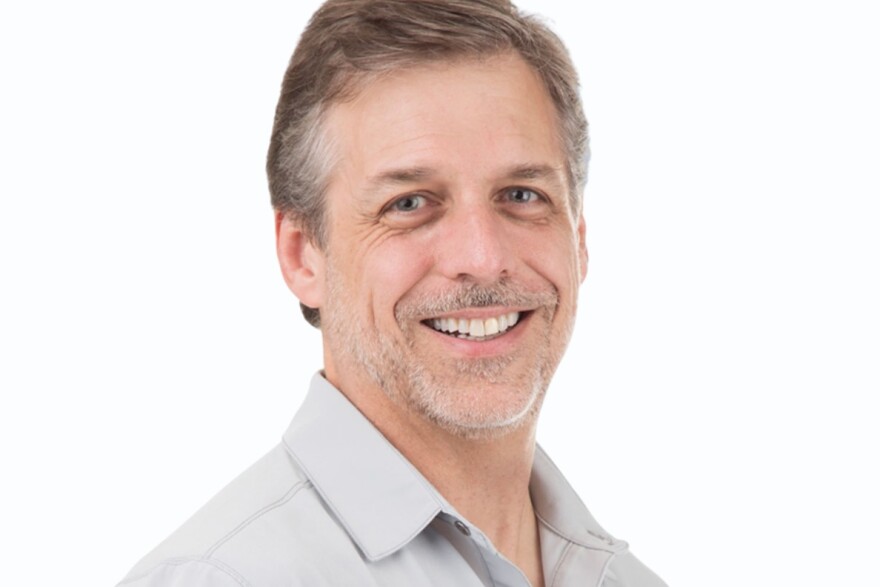 A smiling middle-aged man with short graying hair and a trimmed beard poses against a plain white background. He is wearing a light gray collared shirt and looking slightly to the side.