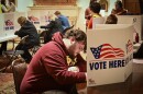 People sit at long tables, bent over while they write. Scattered around them are cardboard dividers that are printed with a waving American flag and read "Vote Here."
