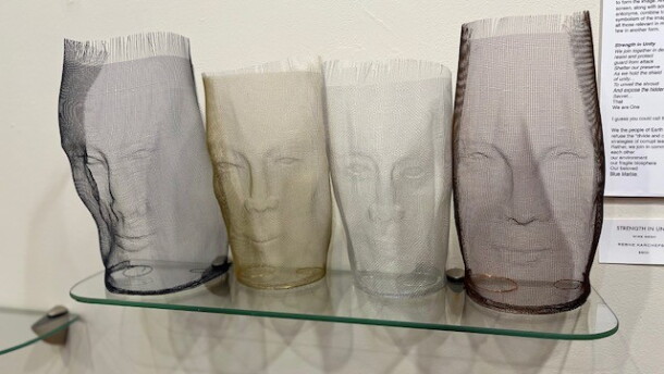 Aluminum wire sculptures of four different heads on a glass shelf and in front of a white wall.