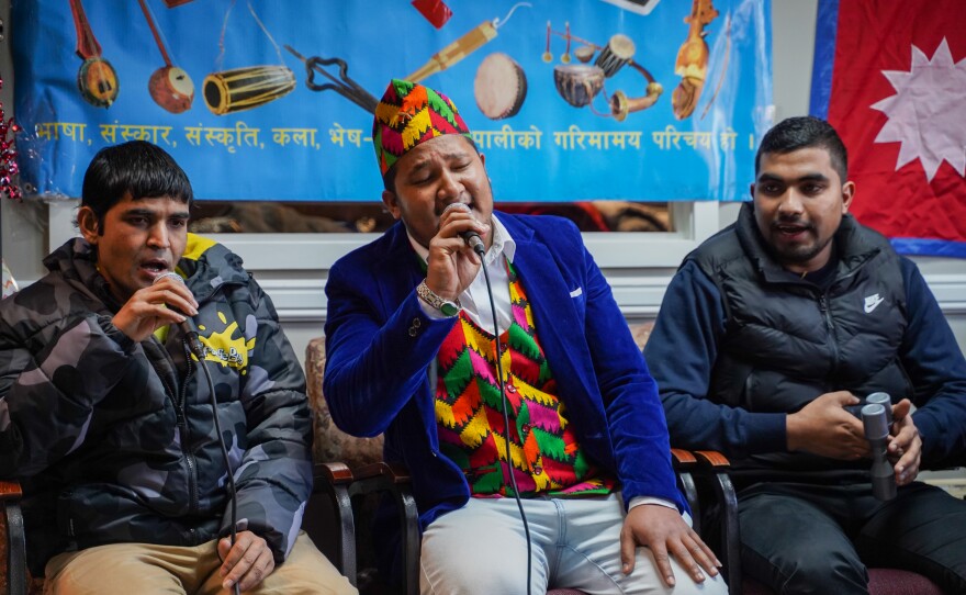 Three seated Nepali men sing, two of them holding microphones.