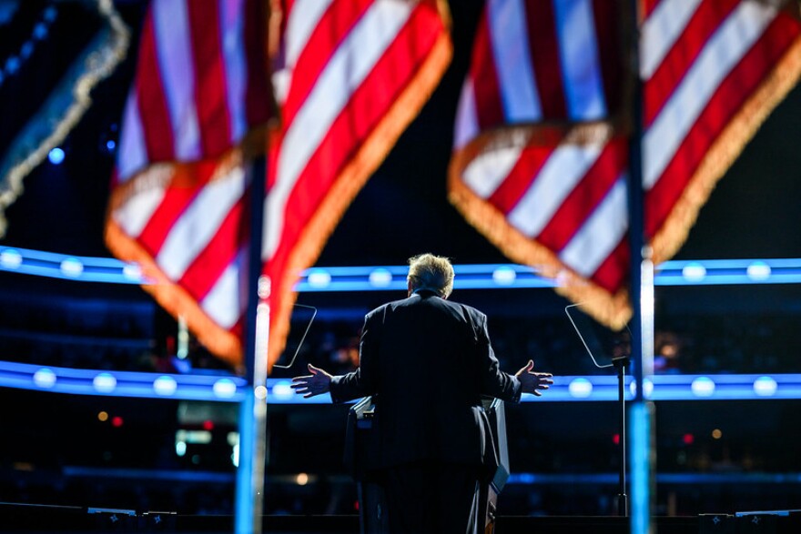 President Donald Trump delivers a speech at the America Business Forum Miami at the Kaseya Center. In the background you can see two blurry United States flags. 