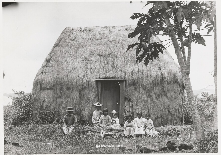 A Hawaiian family sits in front of a grass hut in 1852.