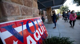 A red, white and blue sign reads "Vote Aqui Here" outside a building where socially distanced people wait to vote.