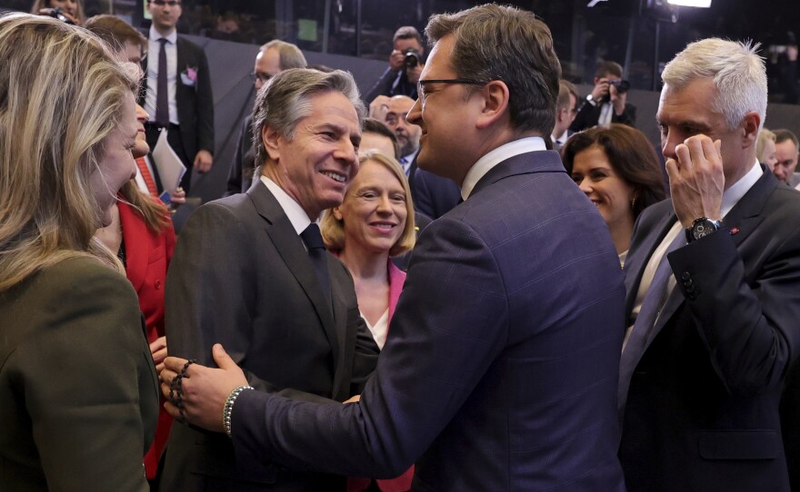 Two men in suits grin while holding each others' arms, as other smiling men and women look on. 