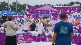 The area is awash with bright white foam, which covers many of the kids pictured in the image. Two adults stand in the foreground, backs to the camera as they watch the children play in the foam inside an area blocked off by bright purple fabric barriers. The fences and matching backdrop all show the name of the area; Foam Fun Zone.