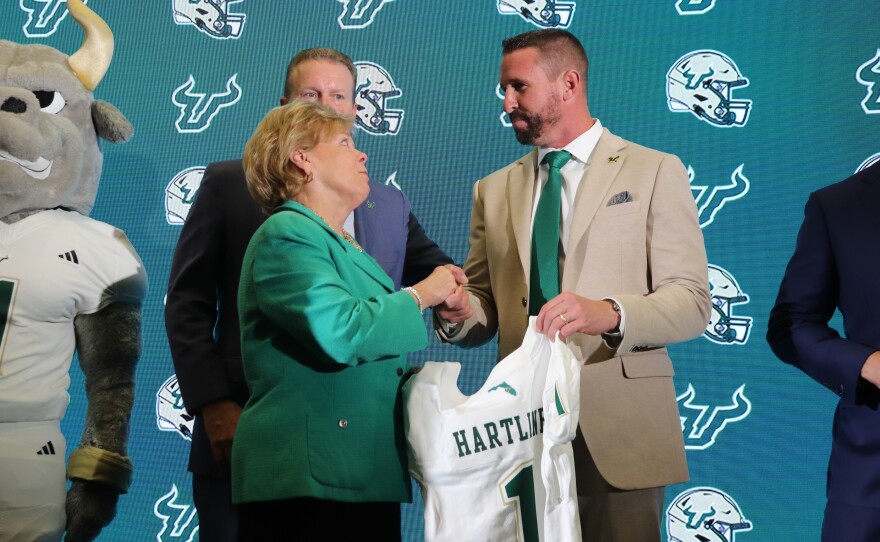 A woman in a green blazer shaking hands with a man in a beige blazer, with his other hand holding a football jersey.