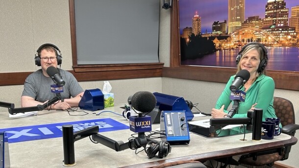 Two smiling people wearing headphones sit at a table in a radio talk studio: A man at left has short brown hair and is wearing glasses and a gray t-shirt; a woman at right has shoulder-length blonde hair and is wearing a green collared shirt, white pants, and white shoes 
