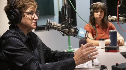 Two women sit inside a radio studio. The one nearest the camera (Sarah Morris) is gesturing and talking. The other woman (Lisa Rodriguez) is listening.