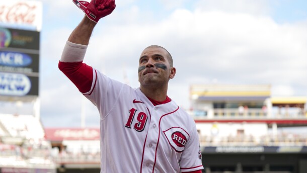 a bald man in a white cincinnati reds uniform looks up and waves at the crowd inside great american ball park