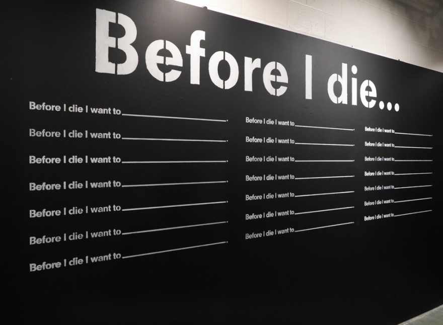 “Before I Die” project consists of a makeshift wall set up for passersby to pick up a piece of chalk and fill in the blank after the phrase “Before I die I want to… .”