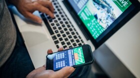 Person's hands holding phone with sports betting app on screen while also typing on laptop with sports game on screen