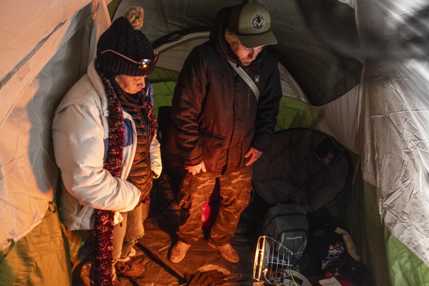 Two people in winter clothing stand inside a tent, looking down a heater fan.