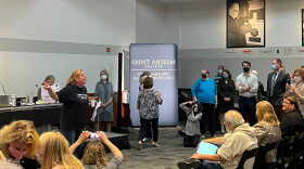 A woman in a black tee-shirt stands in front of a dais, flanked by a crowd. She's gesticulating, as others sit in seats in front of her.