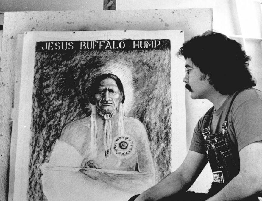 A black and white photograph of a man in overalls with dark longish hair looking at a painting of a Native American man. 