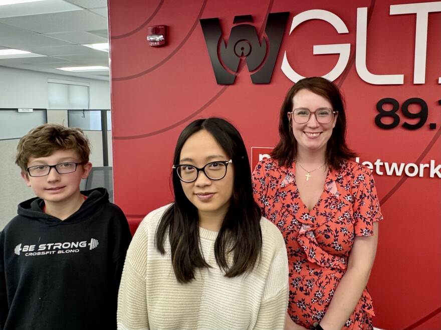 Three people stand in front of a red wall with the WGLT 89.1 radio station logo. On the left is a boy in a black hoodie, in the center is a woman in glasses and a cream sweater, and on the right is a woman in a red floral dress.