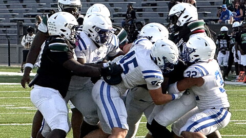football action showing several players in a white uniform with blue strips tackling a player in a black jersey. both teams wearing white helmets. set on a green field with white football lines