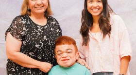 Jeanette, Jasmine and Dominic Henriquez pose for a family photo. Jeanette Henriquez stands on the left in a black short sleeve top with white spots. She has shoulder length strawberry blond hair. Jasmine Henriquez stands on the right in a three-quarter sleeve light colored top. Dominic Henriquez is in front of his mother, Jeanette, and his sister, Jasmine. He has short read hair and is wearing a teal shirt. They stand in from of a white background.