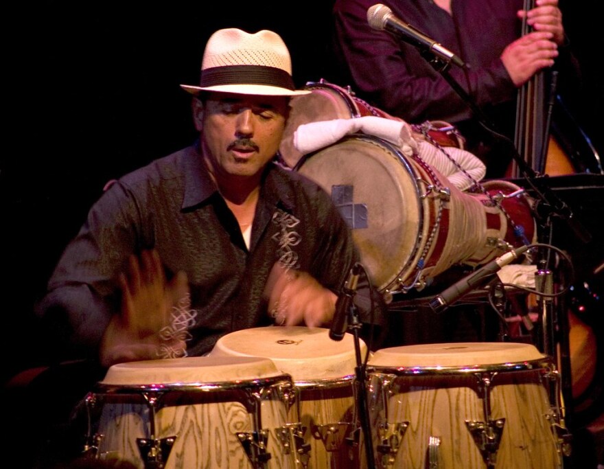 A man in a off-white fedora plays conga drums.