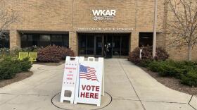The photo shows the front entrance of Michigan State University's Communication Arts & Sciences building labeled with "WKAR PBS NPR Communication Arts & Sciences." There are two signs in front of the building: one that says "REGISTER TO VOTE HERE" and another that says "VOTE HERE." The building is made of brick and has a large glass entrance. There are yellow benches on the side of the building near the entrance. 
