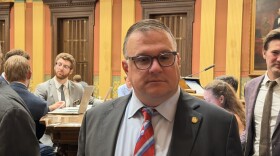 A man wearing a suit with a red, white, and blue striped tie stands in a yellow and orange room where people around him talk, walk, and work on laptops. He has gray hair, glasses, and is looking off from the direction of the camera.