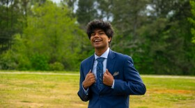 A smiling young man in a blue suit stands outdoors in a grassy field, holding the front of his jacket.