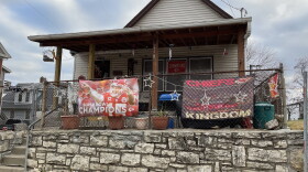 A house is surrounded by a chain link fence. The fence is decorated with banners celebrating the Kansas City football team. Christmas lights hang off the roof and fence.