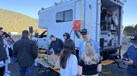 Students and presenters surround a tabletop model of power lines beside a utility truck at an outdoor career fair.