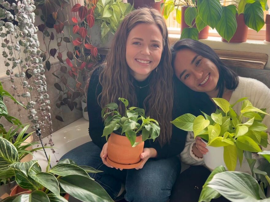 Two women are seated next to each other and smiling while looking toward the camera. The room is covered in hanging and potted plants, and they’re both holding potted plants.