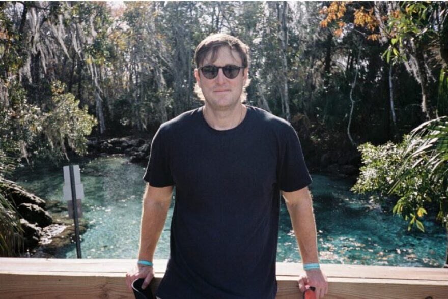 Man with brown hair, dark t-shirt and sunglasses leading back on a wooden railing with a creek behind him and looking into the camera