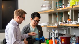 Wyatt Shields, a man in a light colored button down shirt, points to an object that Jin G Lee, a man wearing a gray cardigan sweater and light green T shirt, is holding in his blue-gloved hands. The two men are in a laboratory at the University of Colorado.