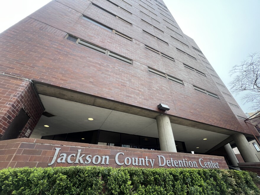 A large, multistory brick building looms over a hedge row. A sign behind the hedges reads "Jackson County Detention Center."
