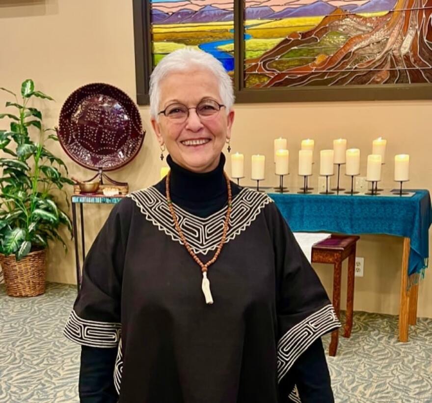 A smiling woman with short white hair wearing glasses, dangling earrings, a long necklace, and a black dress. She is standing in front of a table of candles.
