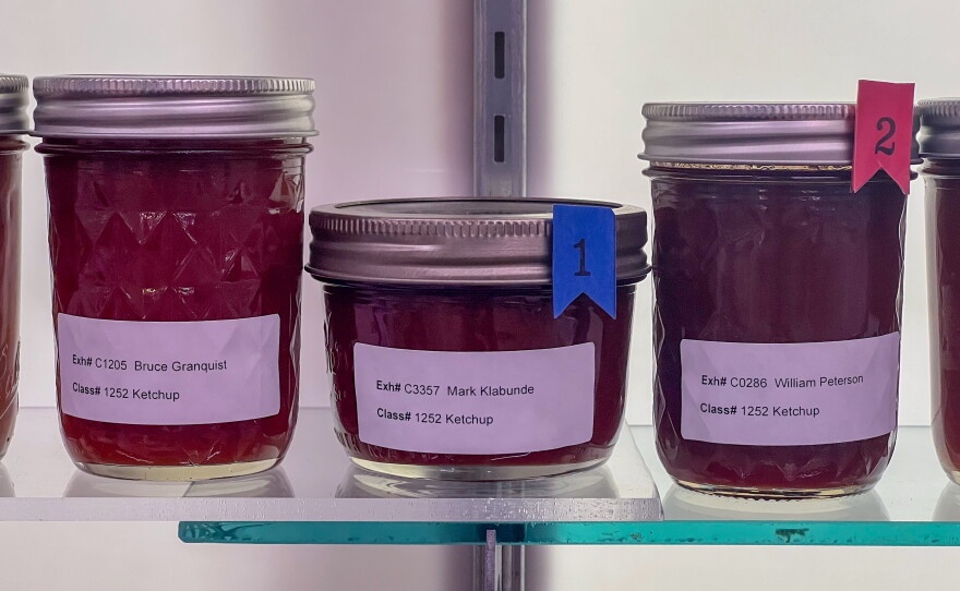 Three jars of homemade ketchup sit on a well-lit glass shelf. 