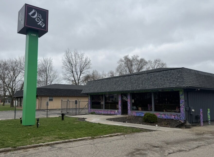 On a cloudy day, a building with a purple swirly paint pattern and a black shingled roof can be seen. In front of the building, a large green sign reads "Doja".