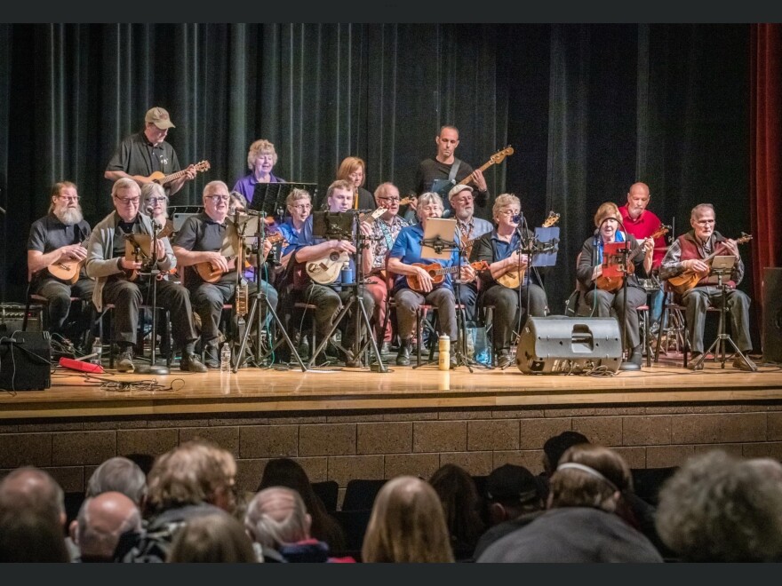 Seventeen people on a stage, most seated and playing ukuleles, in front of a seated audience