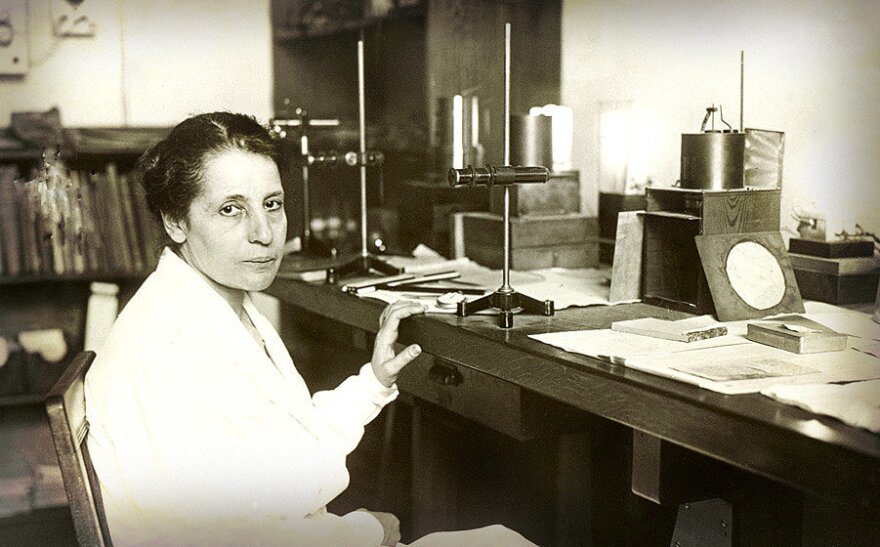 Black-and-white photo of physicist Lise Meitner seated at a laboratory desk, surrounded by scientific instruments and papers, circa 1930.