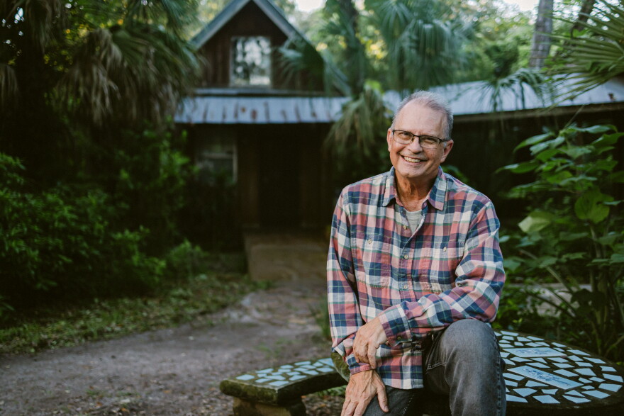 A man in a plaid long sleeved shirt is sitting outdoors on top of a decorative tiled table that's in front of a forrested cabin