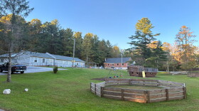 A green mowed yard with a swing-set, a wooden ship with a slide, and a fenced in area, with several building in the background. 