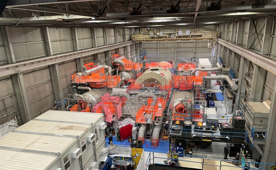 Looking down on the inside of a massive, hangar-like building. The room is filled with metal equipment, including the Palisades turbine, which sits partially dissassembled. Orange netting covers much of the area around the building.