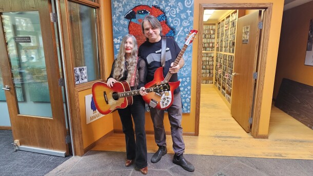 A man and woman holding guitars in front of a painting. The woman has an acoustic and the man has a electric guitar.