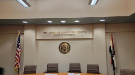 Brown high-backed chairs sit around a tan panel desk with microphones. The American flag sits to the left of the seal of the state of Missouri and the Missouri flag to the right. The words the grand jury of St., Louis County, state of Missouri, are written on the wall above the seal.