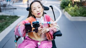 a woman in brightly colored, loose-fitting clothes, with a half-smile and brightly colored lipstick, sitting in a wheelchair with a breathing tube going from her neck to the back of the chair