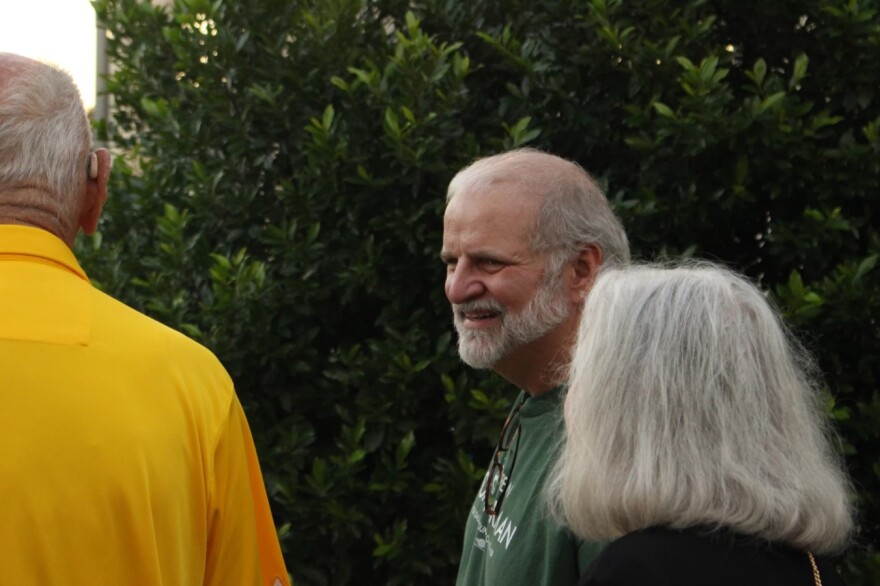 Gray haired man in green shirt smiling, looking at a man in yellow shirt. Behind him ins a large bush or tree.