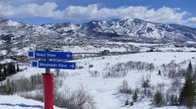 On a mountain in Steamboat Springs, two blue signs sit on top of a pole wrapped with red padding. The blue sign on top reads "Town View" with an arrow pointing to the left. The blue sign on the bottom reads "Mountain View" with an arrow pointing to the right. Snow-covered mountains and a blue sky are in the background